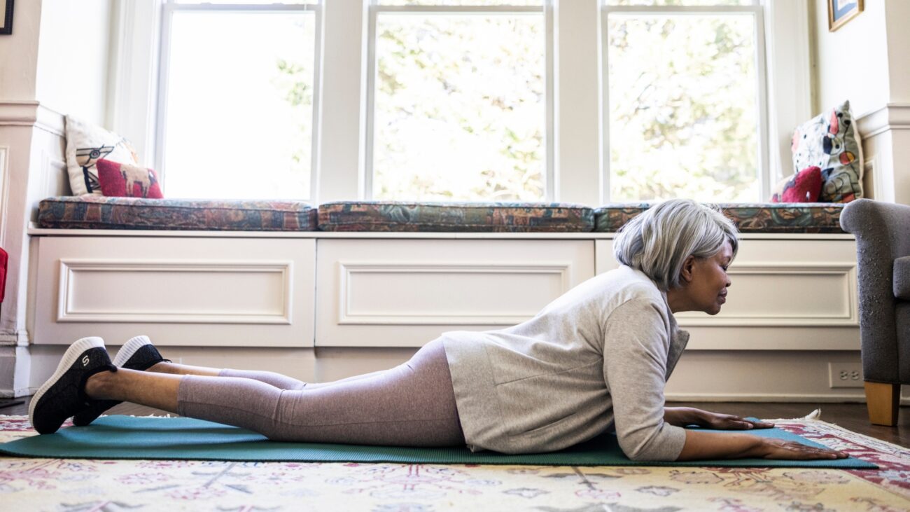 older woman lying on her front and forearms on an exercise mat in a living room setting with big windows behind her