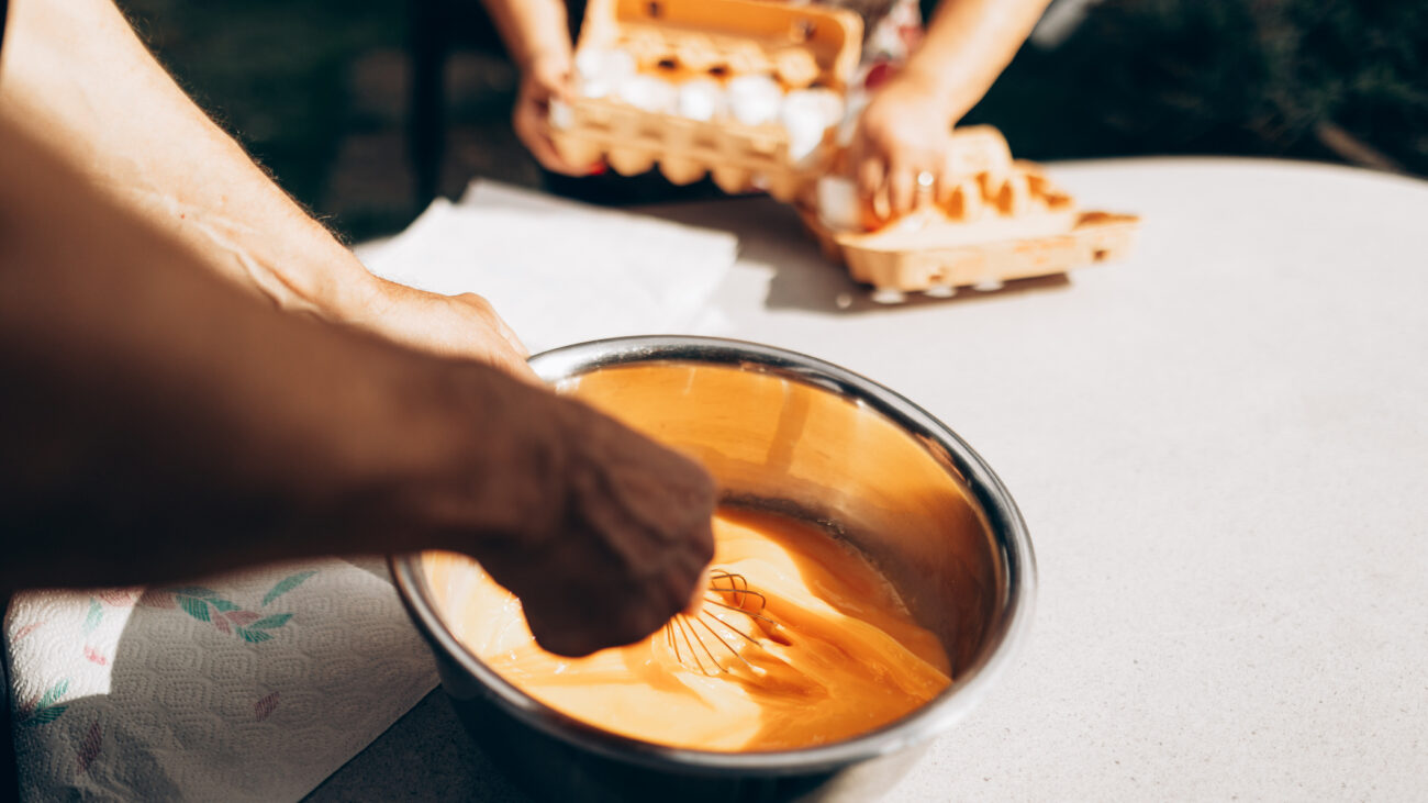 Hands beating eggs in a metal bowl with a whisk. Another person's hands are in the backgrounds with two large cartons of eggs
