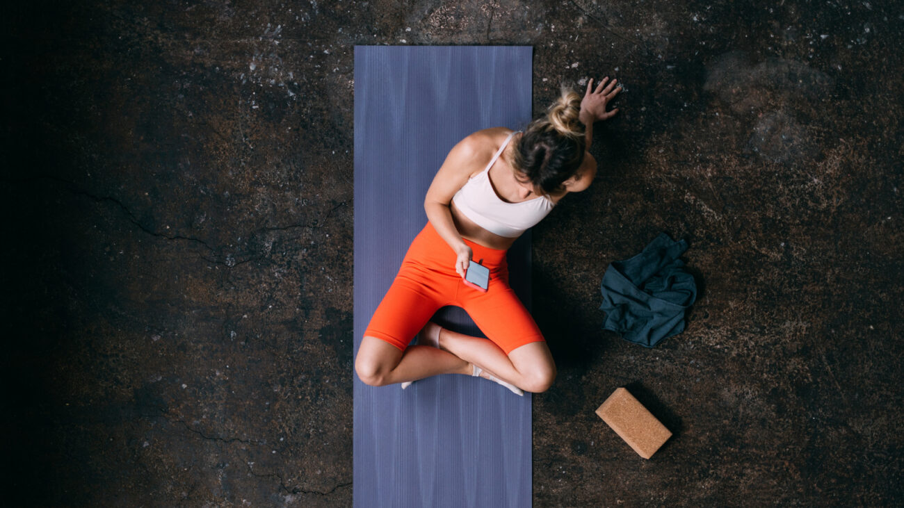 Woman seen from above sitting on yoga mat looking at smartphone with a cork yoga block next to her