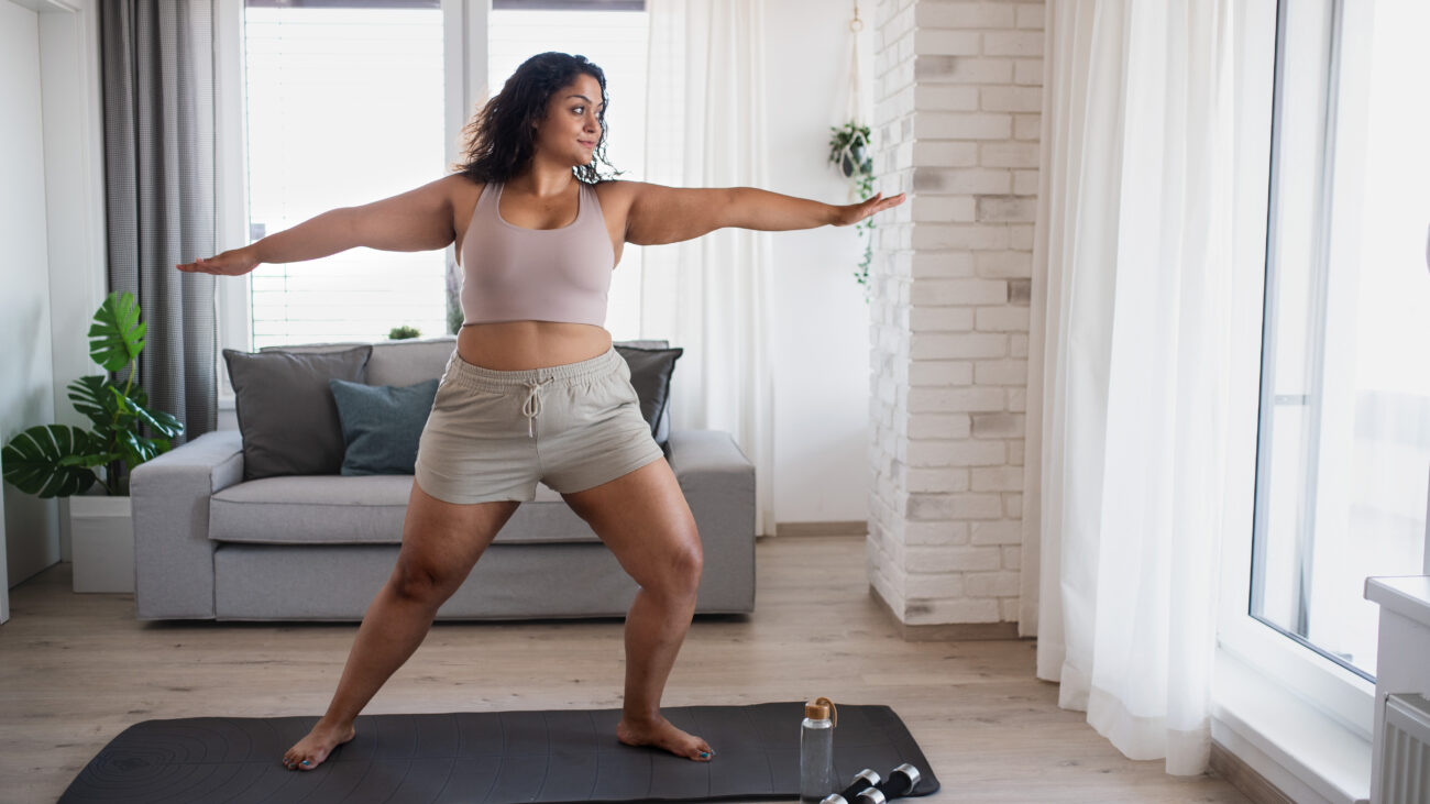 A woman performs a lunge in a yoga routine. She is in a living room, practicing on a yoga mat, with a couch and plant behind her.