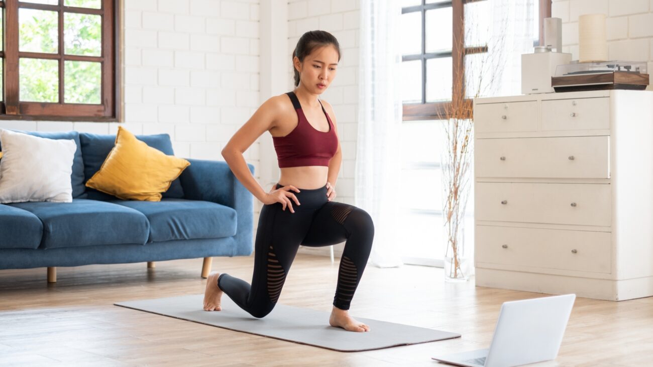 woman in black leggings and red crop top performing a lunge on an exercise mat in front of a laptop on wooden floor. there's a navy sofa and windows behind her and a white chest of drawers next to her.