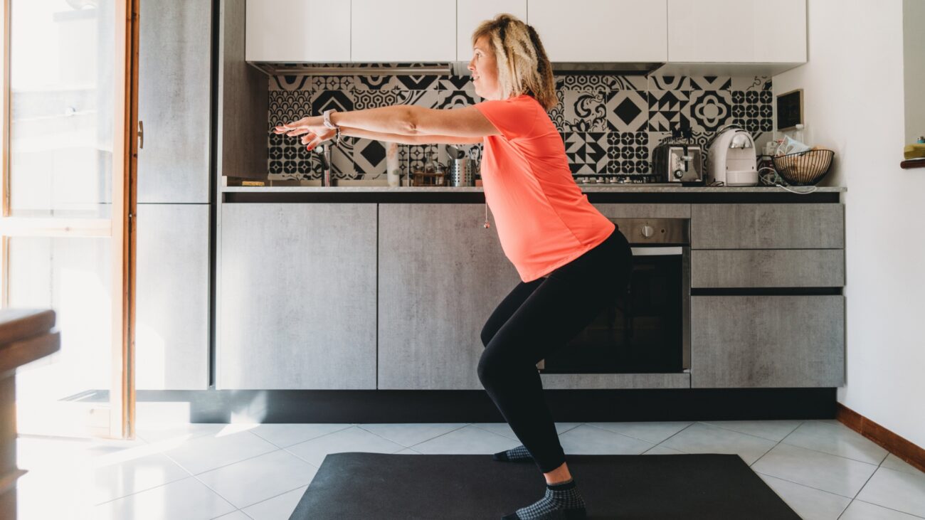 woman performing a squat in her kitchen, sideways to the camera. she's wearing a coral coloured tshirt and black leggings, standing on a black exercise mat in front of some silver kitchen counters.