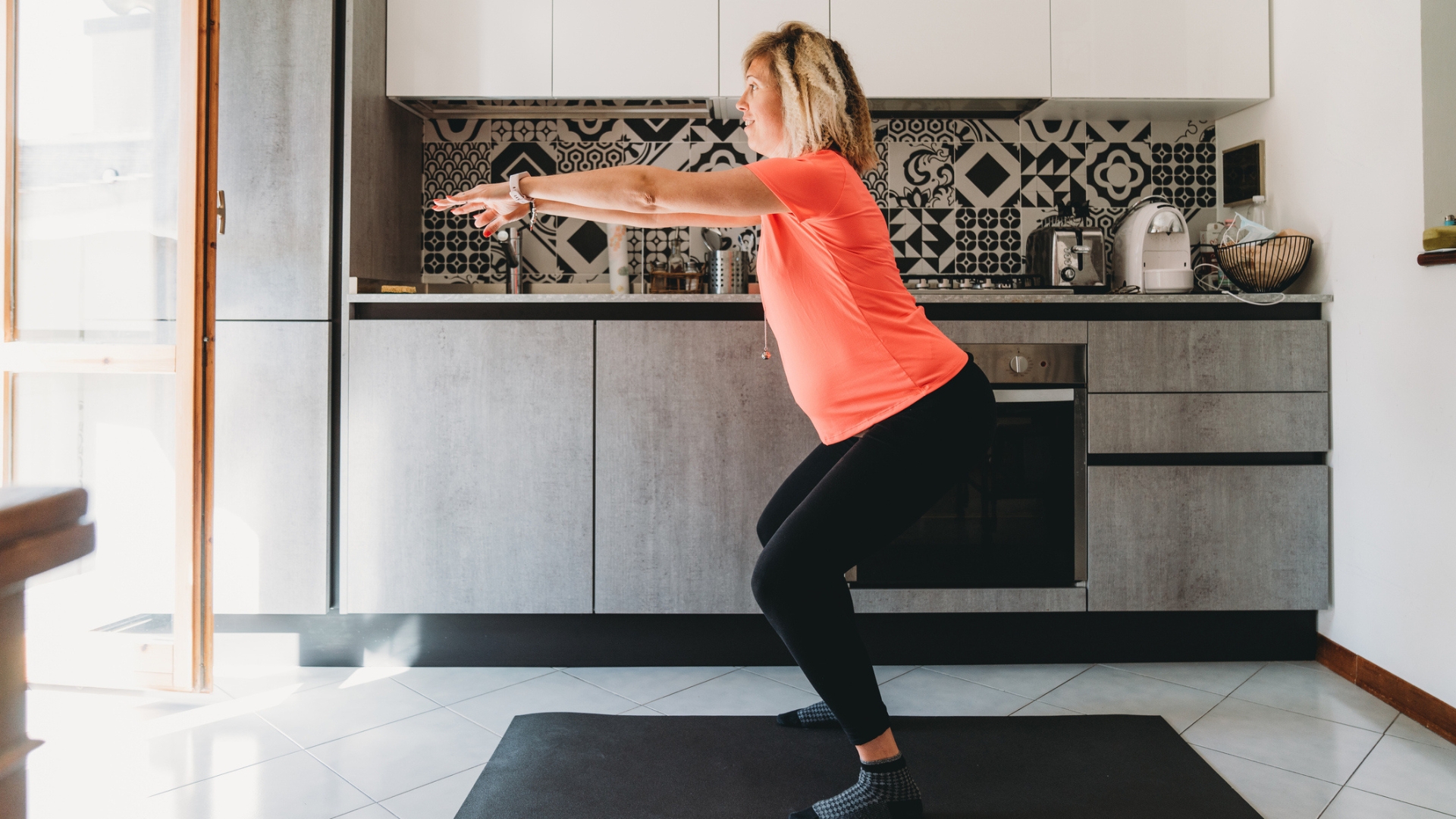 woman performing a squat in her kitchen, sideways to the camera. she's wearing a coral coloured tshirt and black leggings, standing on a black exercise mat in front of some silver kitchen counters.