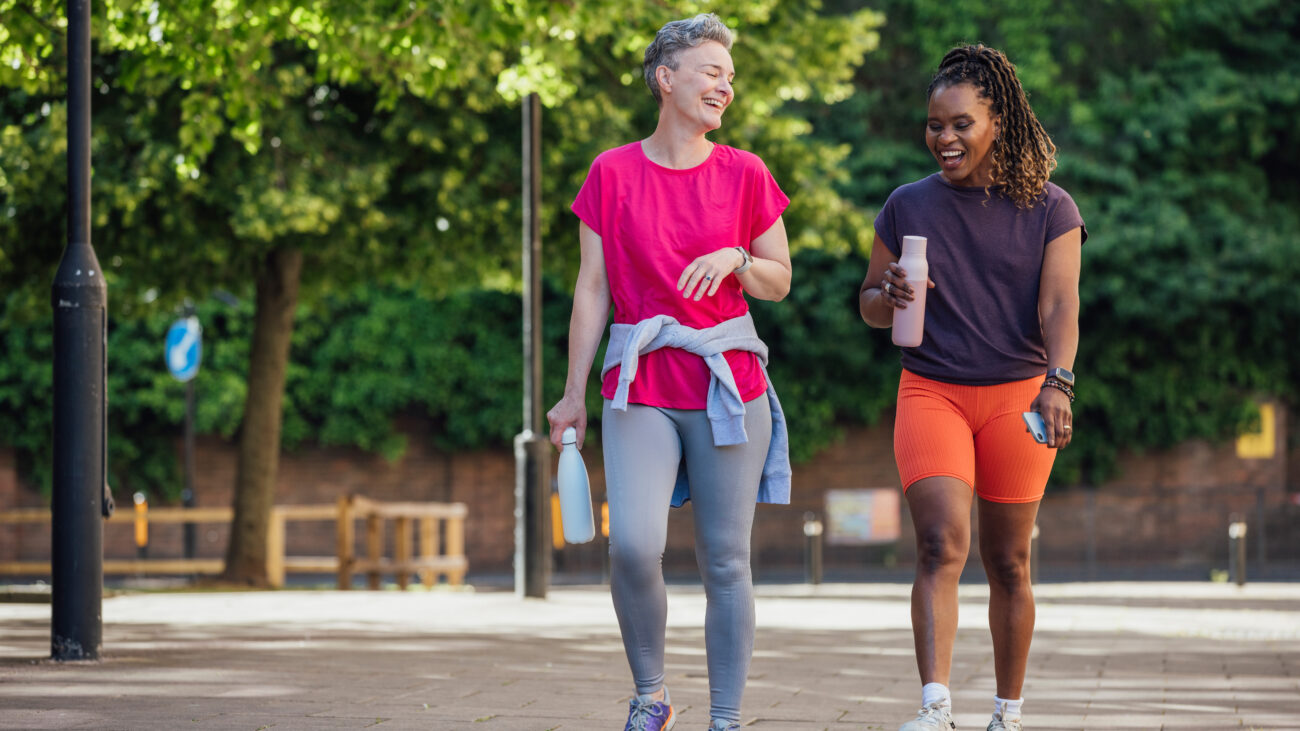 Two women walk along a paved path outside, laughing and holding water bottles. Behind them we see leafy trees and lamposts.