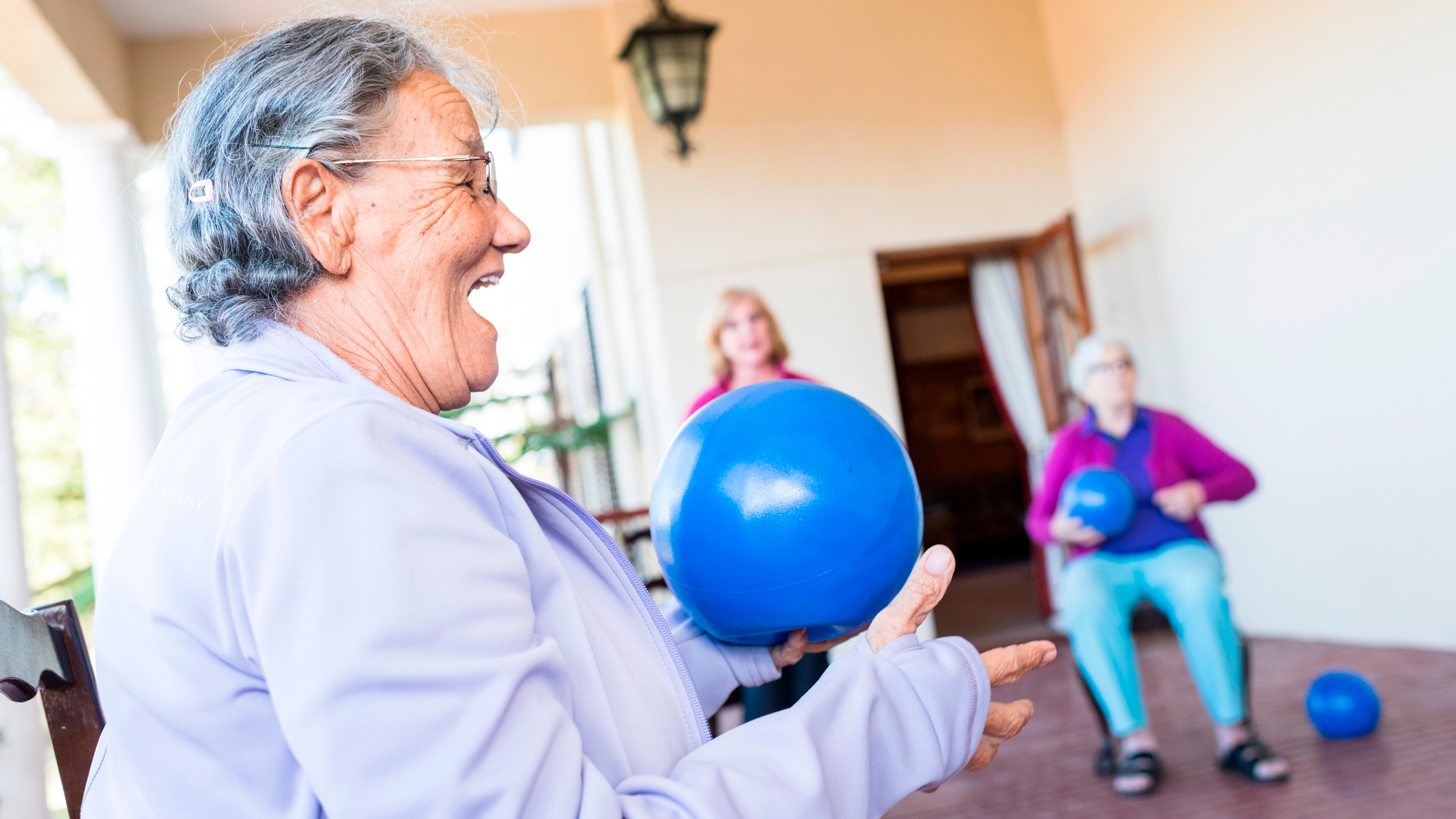 Senior woman sitting in a chair holding a blue Pilates ball in her hand. Her mouth is open in a joyful expression. Other women in chairs holding balls are in the background.
