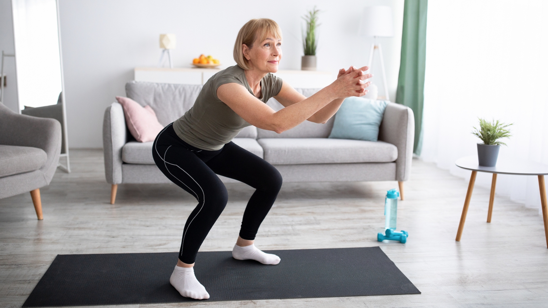 Woman performing a shallow squat on a black exercise mat in a living room setting. there's a sofa behind her.