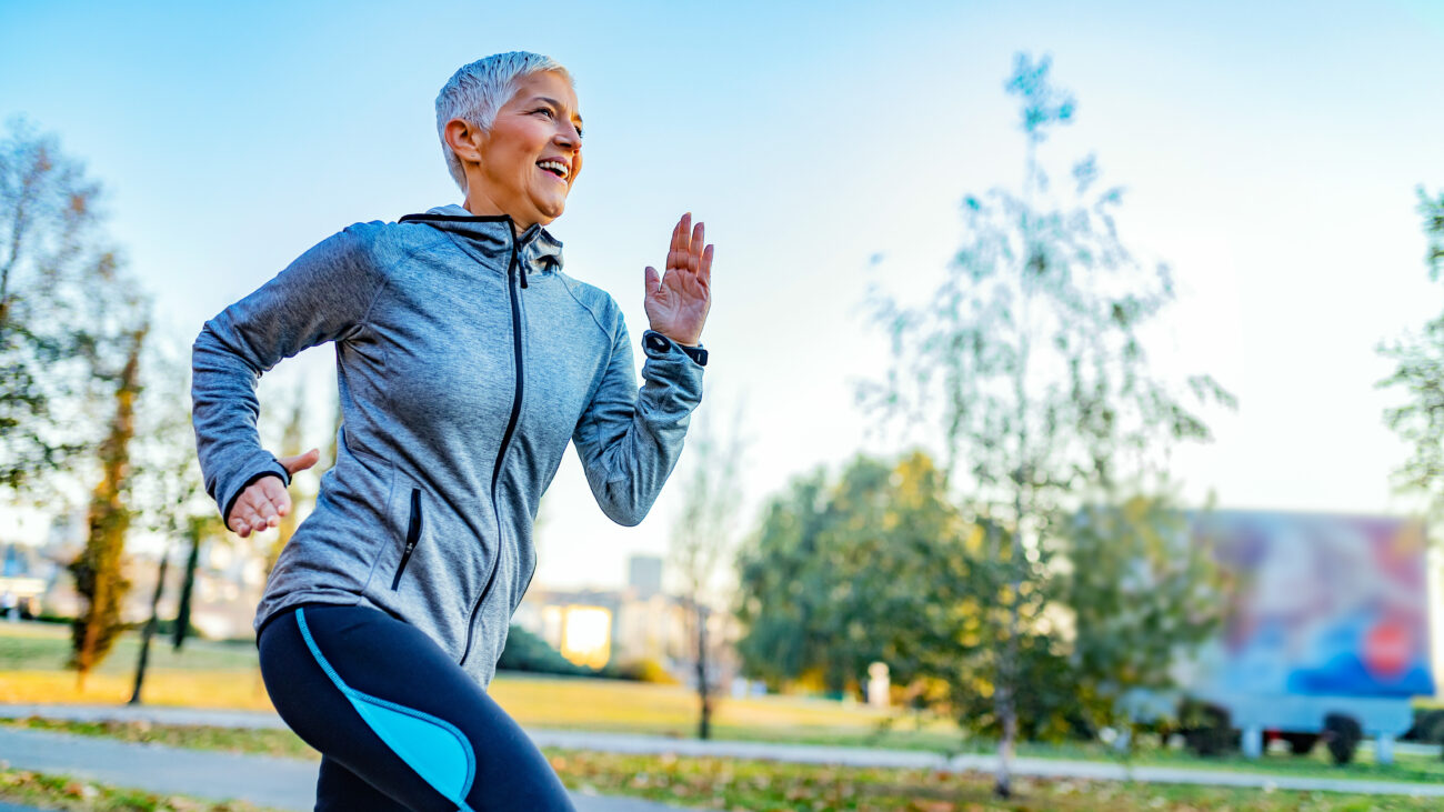 A woman in a sports jacket and leggings smiles as she runs through a park.
