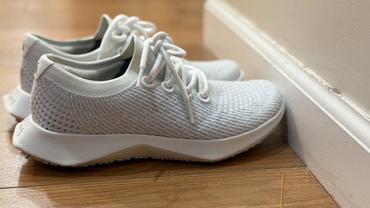 White and grey AllBirds Tree Dasher 2 sneakers on a wooden floor lined up against a white wall.