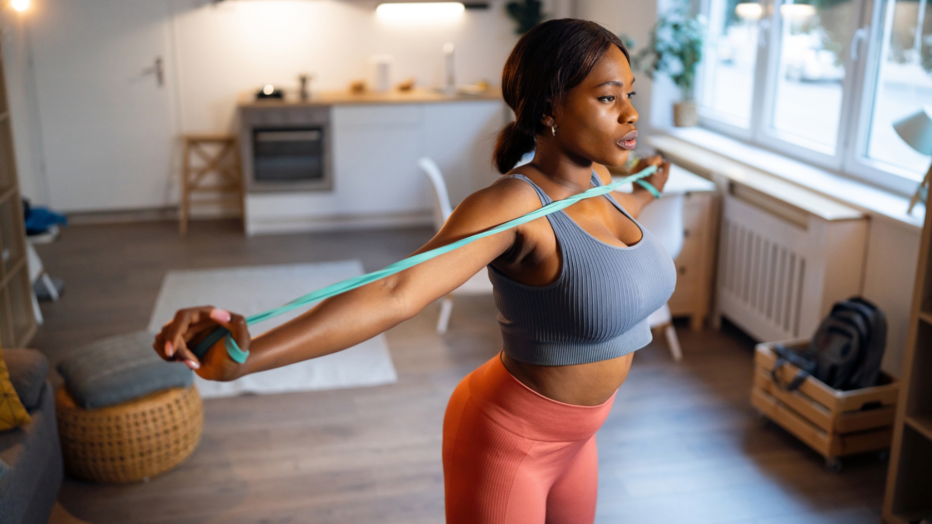 woman holding the ends of a resistance band in each hand and pulling them apart at chest height. she's in a living room setting looking away from the camera.