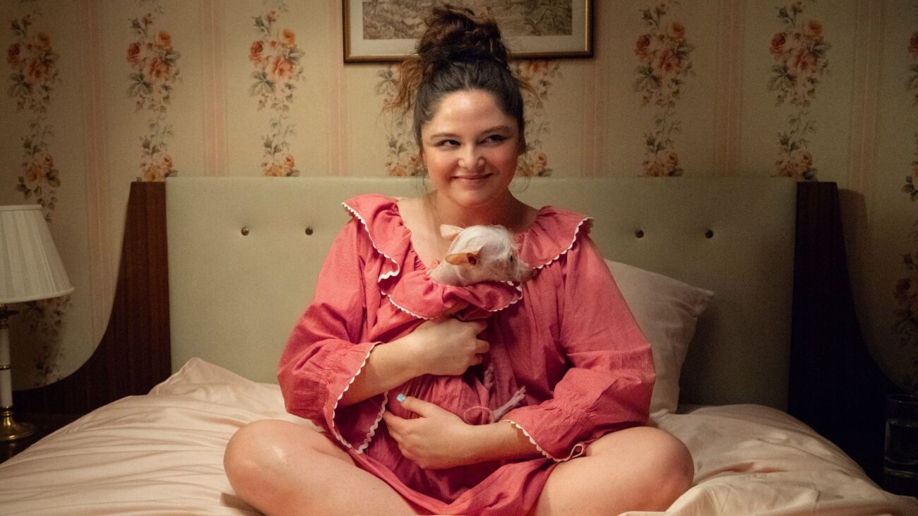 Megan Stalter sits on a bed holding a hairless dog wearing the same pink dress with white trim