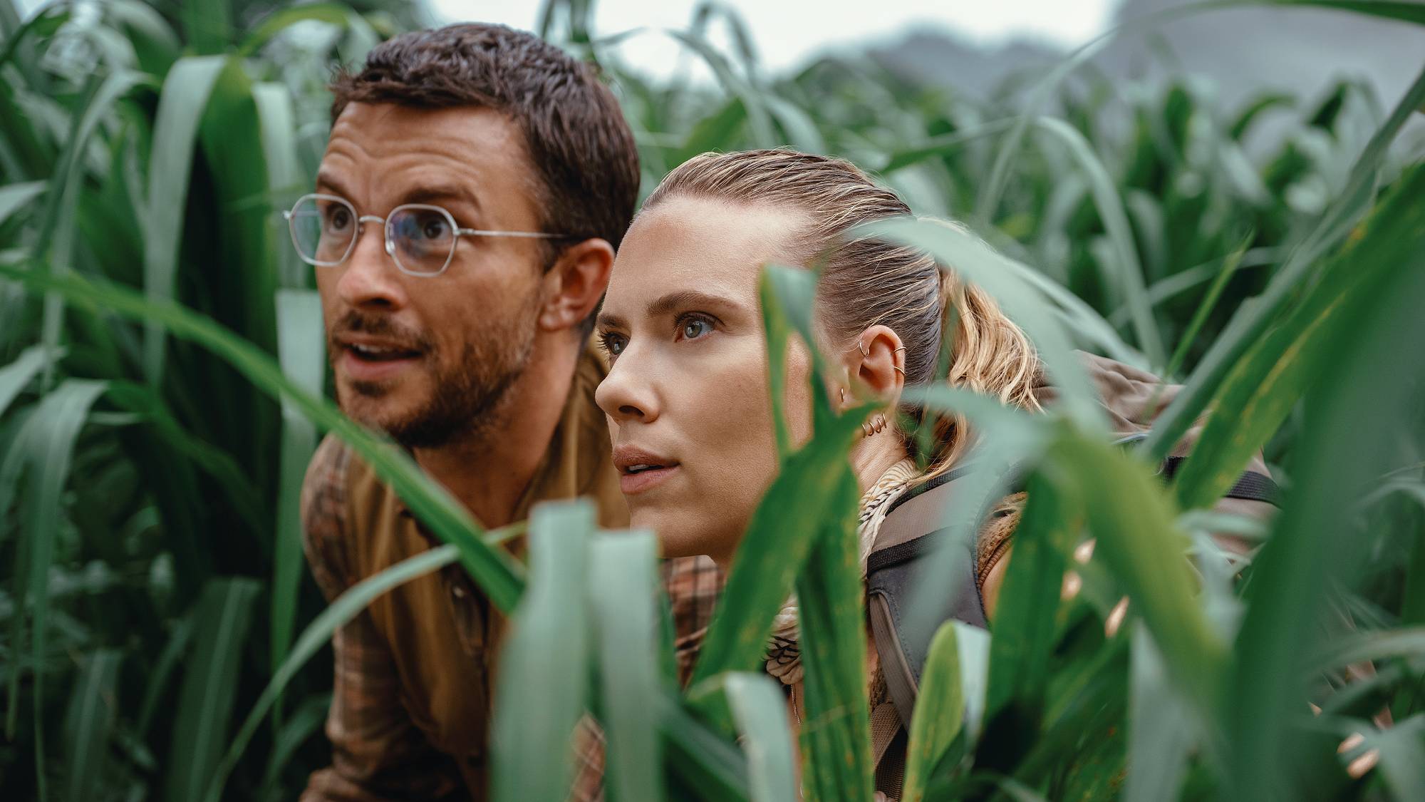 L-R: Jonathan Bailey as Dr. Henry Loomis and Scarlett Johansson as Zora Bennett peering through the undergrowth in a scene from "Jurassic World Rebirth"