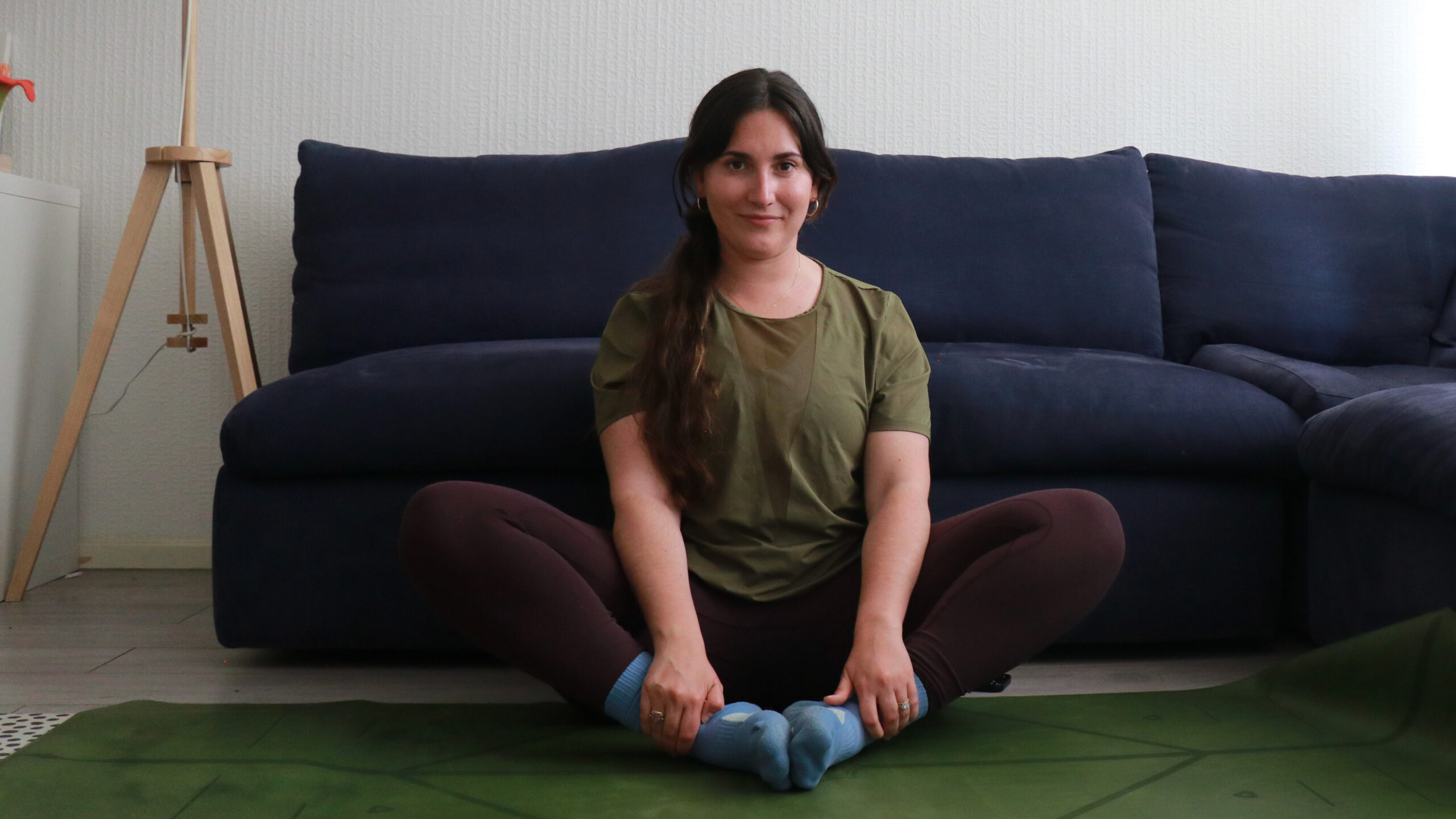 Marina Leiva García performs a butterfly pose at home. She is sitting with her knees bent and the soles of her feet together. Her knees are pointing upwards at a 45° angle. Behind her is a couch.