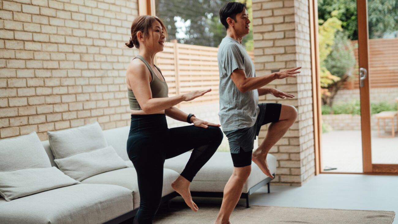 woman and man performing high knees exercise in a living room setting, looking sideways to the camera with a white corner sofa behind them and windows and glass door on the other side
