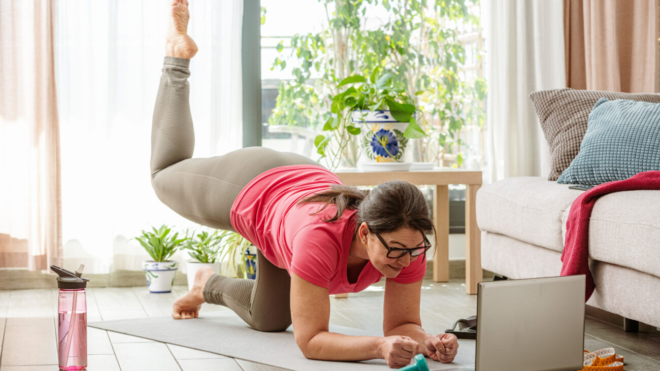A woman practices Pilates at home in her living room on a mat. She is balancing, close to the ground, on her forearms and her bent left knee. Her right leg is bent at 90&deg; and elevated behind her. In the room we see a wtaer bottle, a couch, a plant and a coffee table. A laptop is open in front of her.
