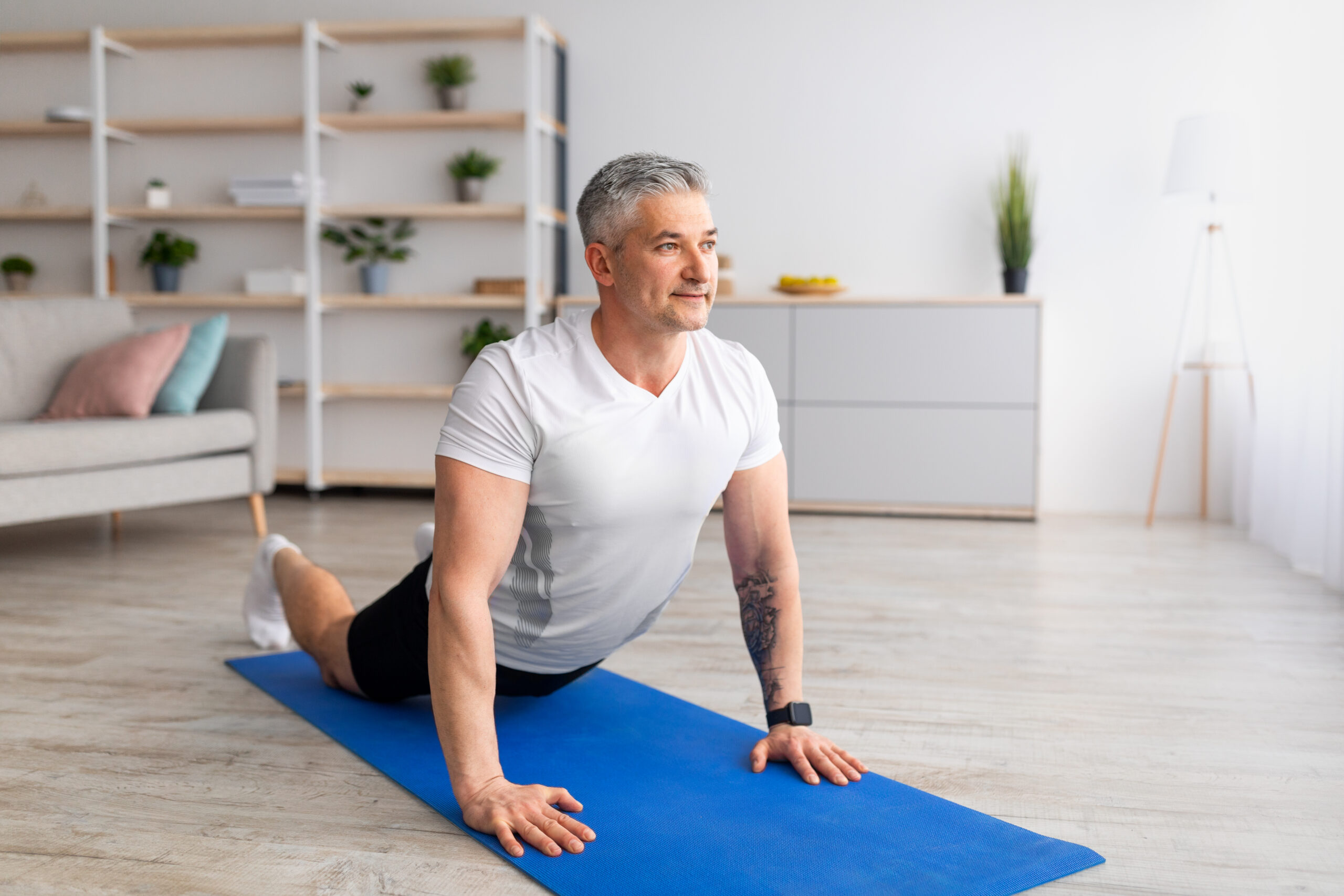 A man practices cobra pose at home on a yoga mat. He is on his front, with his legs long on the mat and his torso held upright by straight arms. Behind him we see a couch and shelves with plants.
