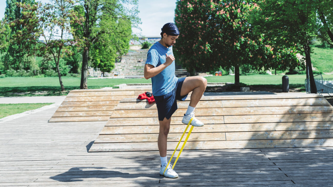 A man performs a resistance band exercise in a park outdoors. He is standing, with the band looped around his feet, and one knee bent so that he is in a marching pose. Behind him we see a long path, steps and trees.