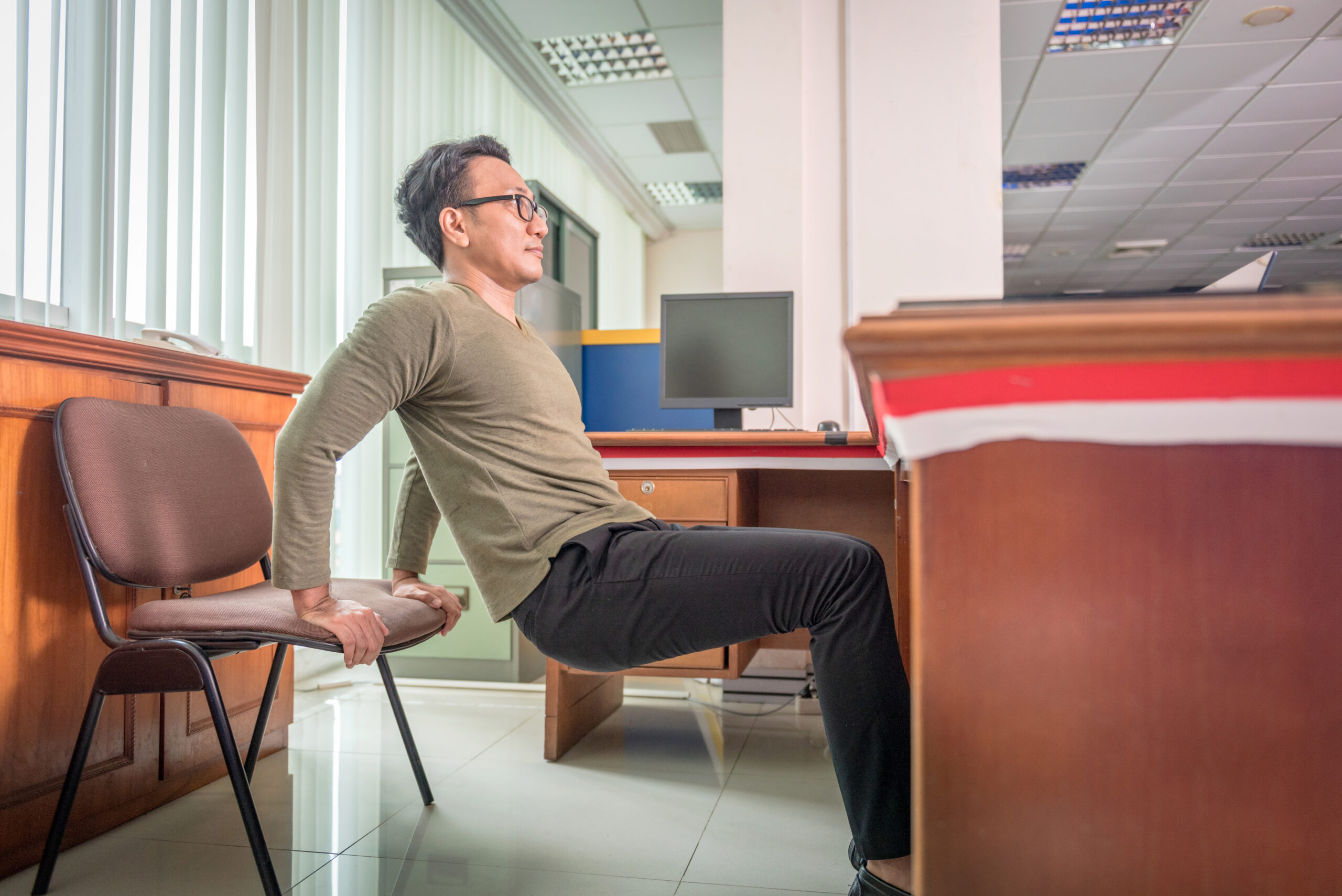A man practices triceps dips at his desk on a chair. He has pushed the chair back away from the desk; his hands are on the edge of the chair, his feet placed far in front and his knees are bent. In front of him is a wooden desk.