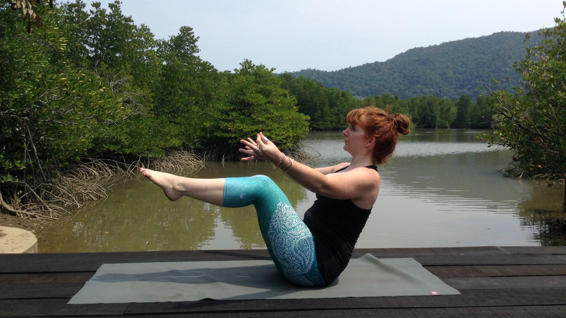 Yoga teacher Alison Reaume in boat pose. She is on a wooden platform with a lake and trees in the background