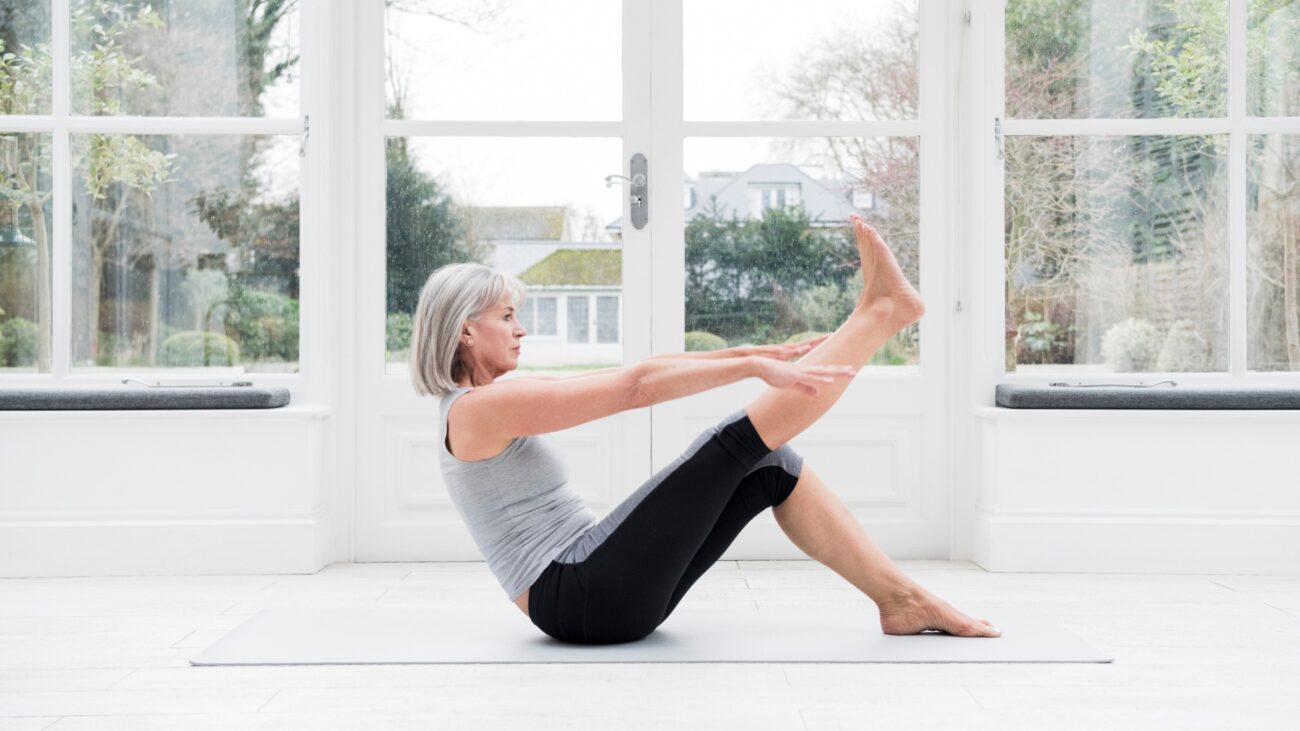 Woman in her 60s with grey bobbed hair stretching leg at home in front of windows overlooking a garden.