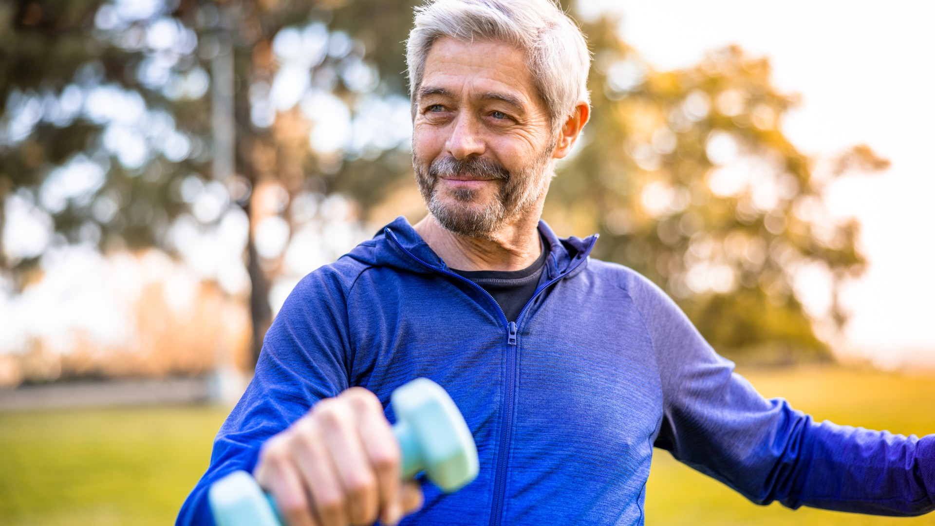 Man over 60 wearing blue tracksuit top holding a dumbbell during workout outdoors