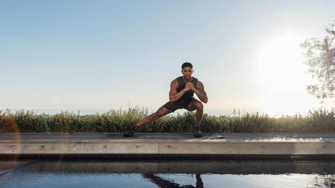 Man demonstrates side lunge exercise outside, he is reflected in a body of water in front of you. There is grass and blue sky behind him.