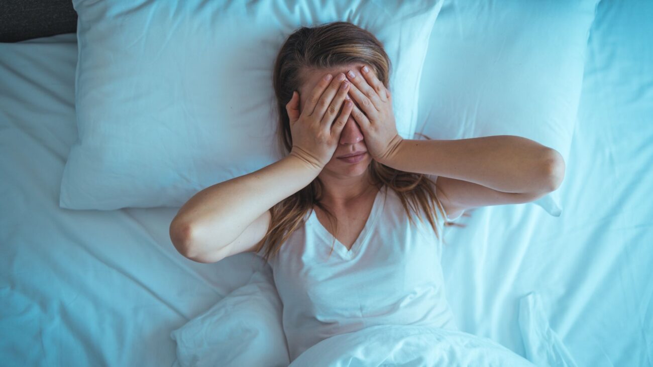 A woman lies in bed covering her eyes with her hands as she struggles to sleep.