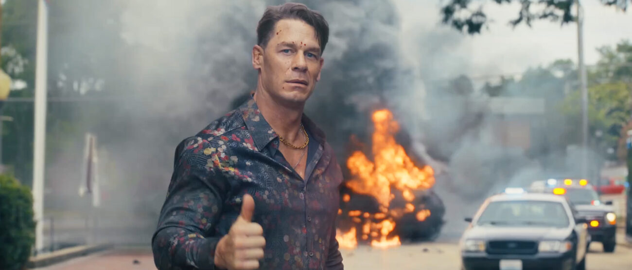John Cena's Chris Smith smiling and giving a thumbs up as a car explodes behind him in Peacemaker season 2