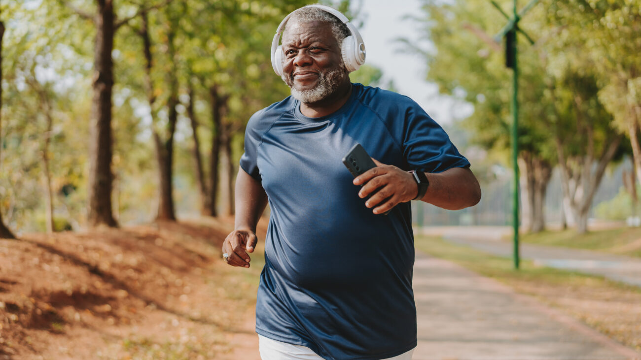 A man runs in a park, wearing a sports t-shirt and a fitness tracker. He has overear headphones on and holds his phone in his hand. Behind him we see a long row of leafy trees.