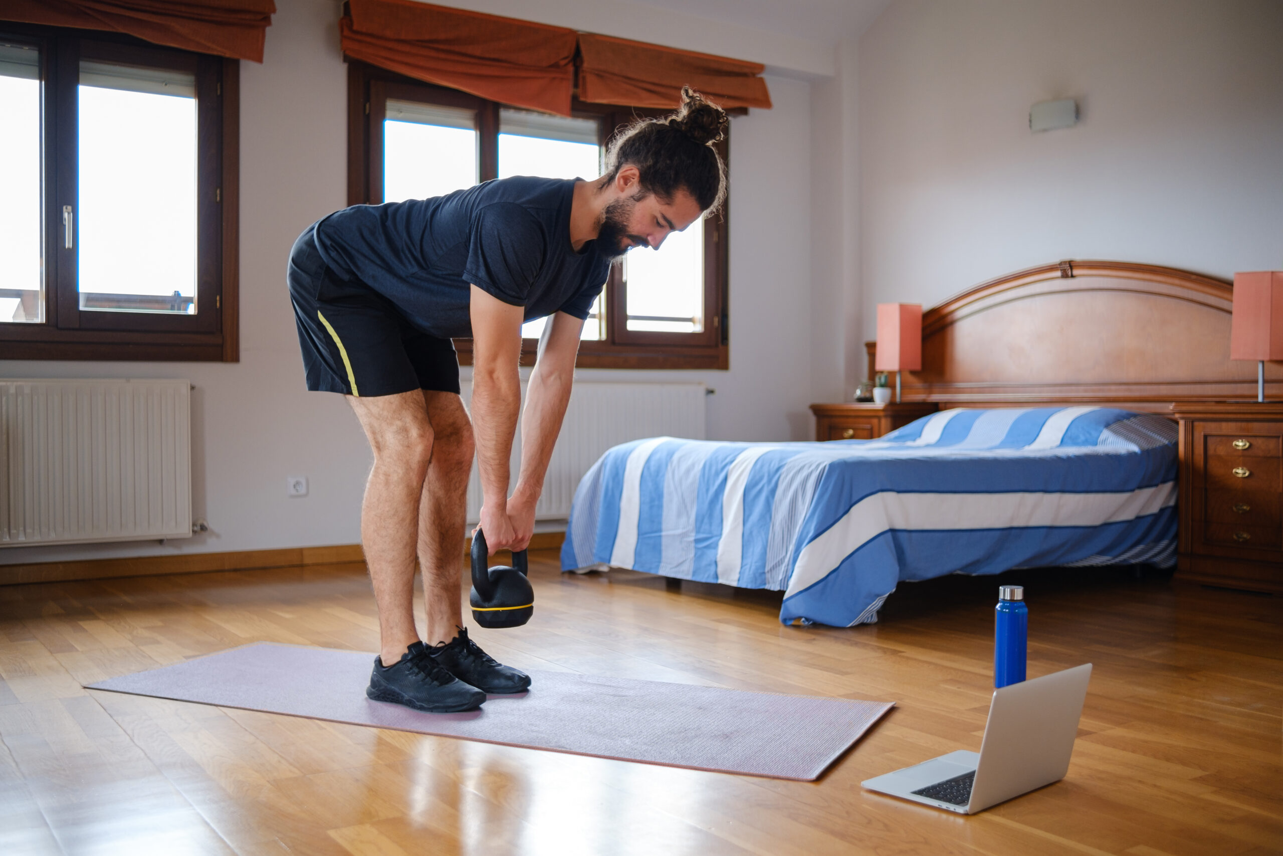 A man performs a deadlift with a kettlebell at home in his bedroom. He is stood with feet together, hips hinging forward so his torso leans forward, and a kettlebell held in his hands at about shin height. We see a bed behind him and a laptop in front.