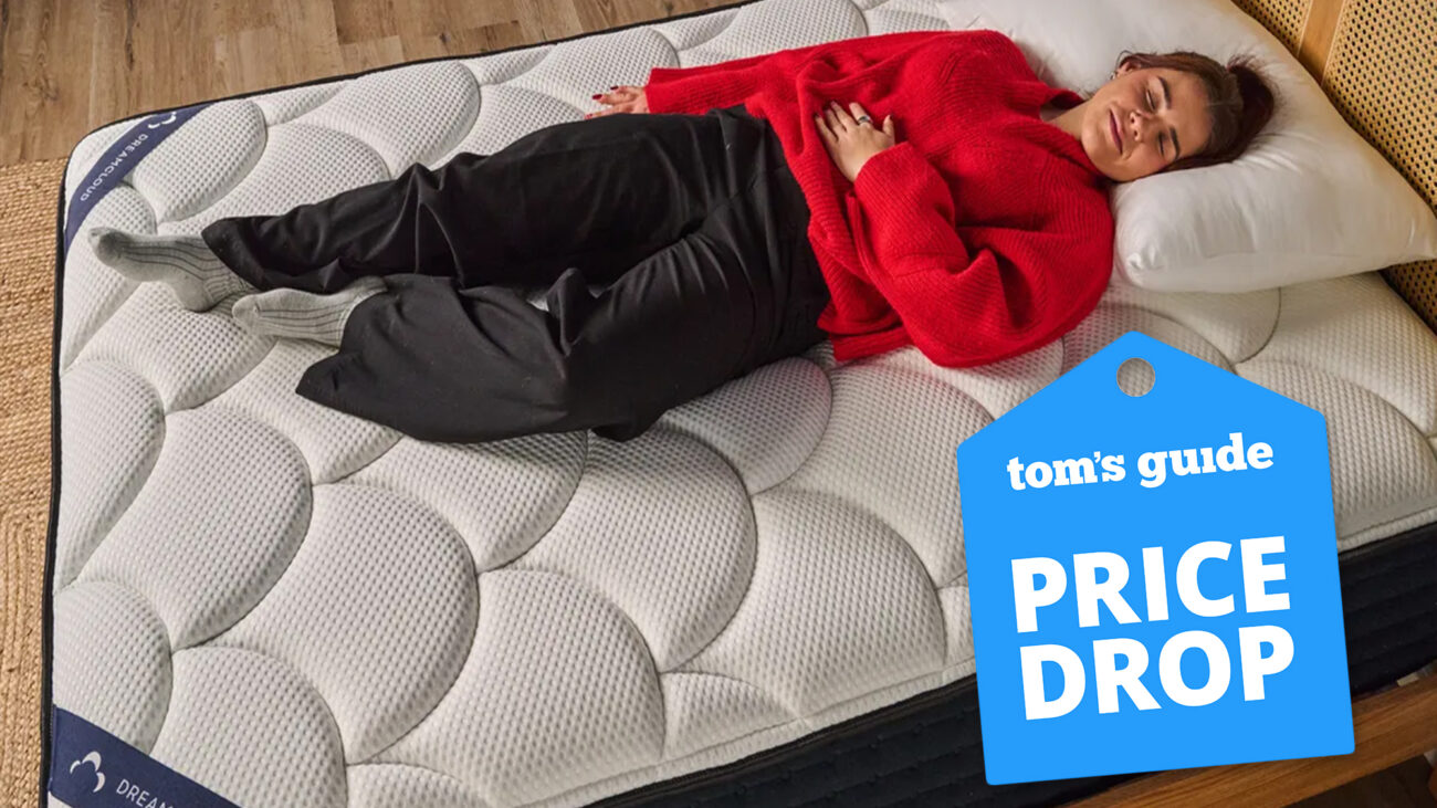 Tom's Guide Sleep Writer Eve Davies lies on her back on the DreamCloud Classic Hybrid Mattress In A Box, dressed in a cozy red top and comfy black pants. A blue Tom' Guide price drop sales badge is overlaid on the bottom left hand corner