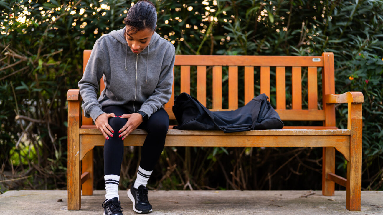 A woman on a park bench wearing workout clothes sits and gently holds her right knee with both hands.