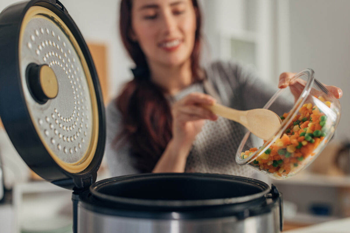 Woman using a wooden spoon to empty a glass bowl of chopped veg into slow cooker