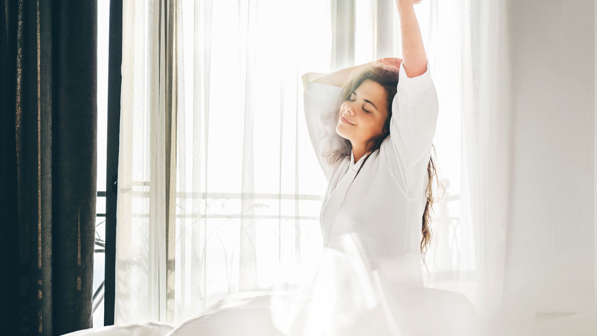 A woman wearing white stretches and smiles as she wakes up in front sunlight streaming in through a window.