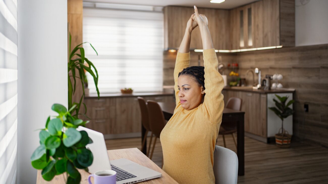 woman in a yellow top sitting in front of a laptop with arms overhead stretching. there's a brown kitchen behind her