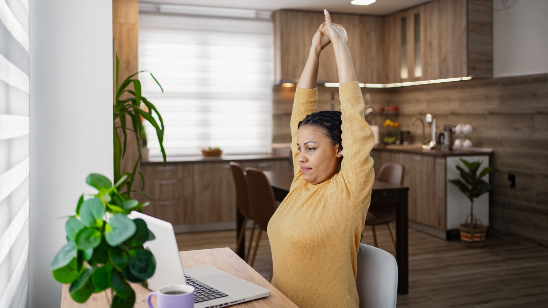 woman in a yellow top sitting in front of a laptop with arms overhead stretching. there's a brown kitchen behind her
