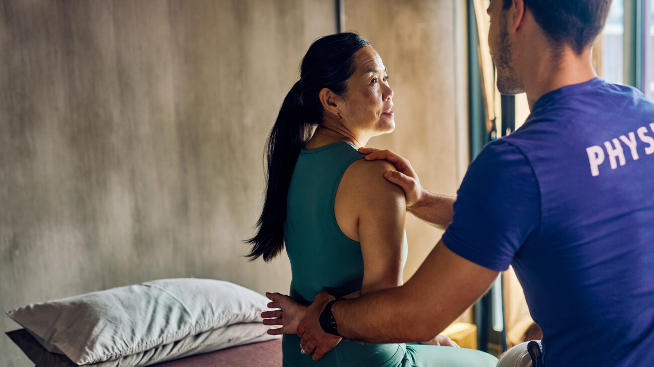 Woman sitting on treatment bed talking to physio who has one hand on her shoulder and one hand on her wrist which is behind her back