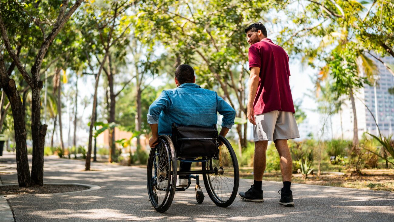 man walking outside with someone in a wheelchair on a path with trees all around in a park setting.