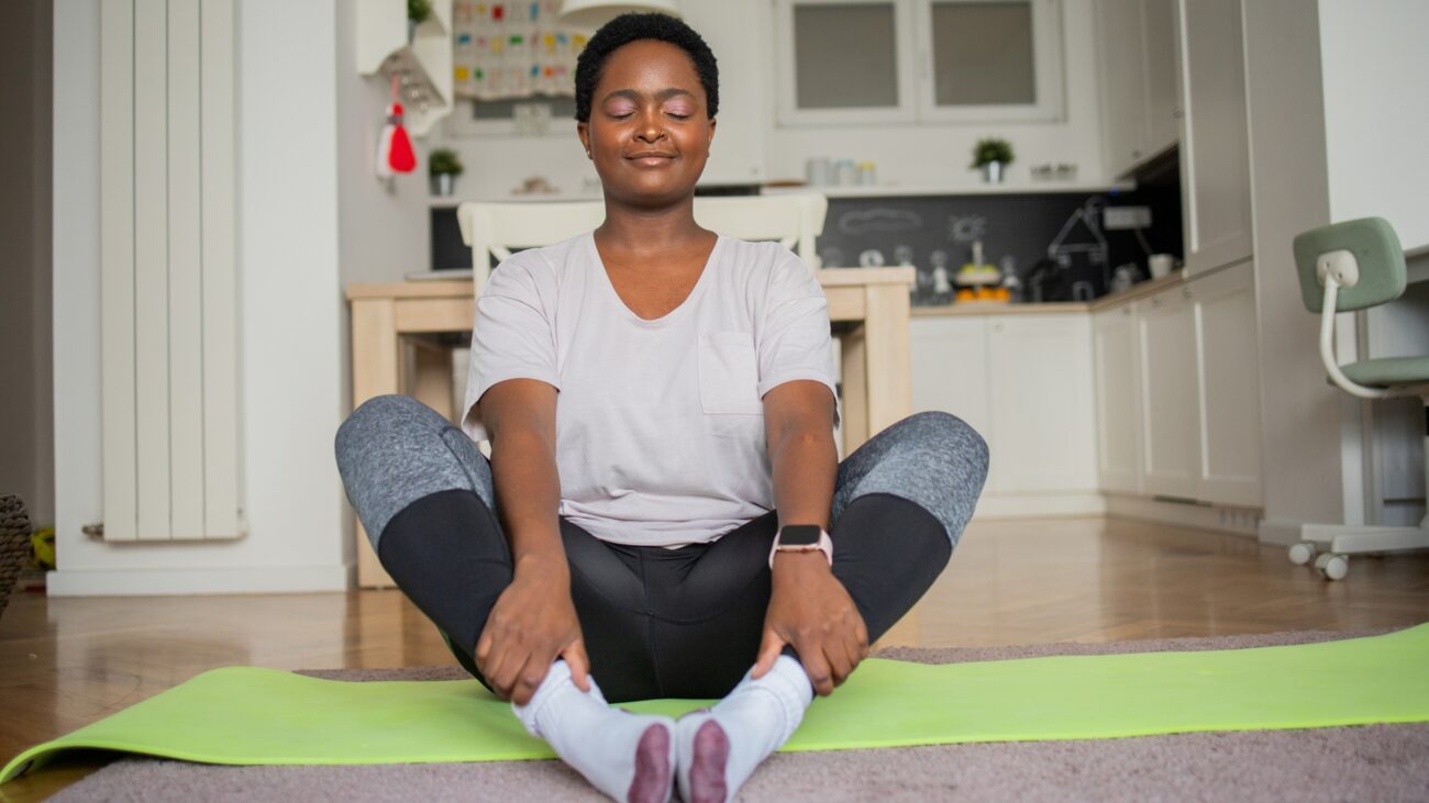 woman wearing a white tshirt and leggings sitting on the floor on a green yoga mat in butterfly pose, facing the camera with eyes closed. there's a kitchen behind her.