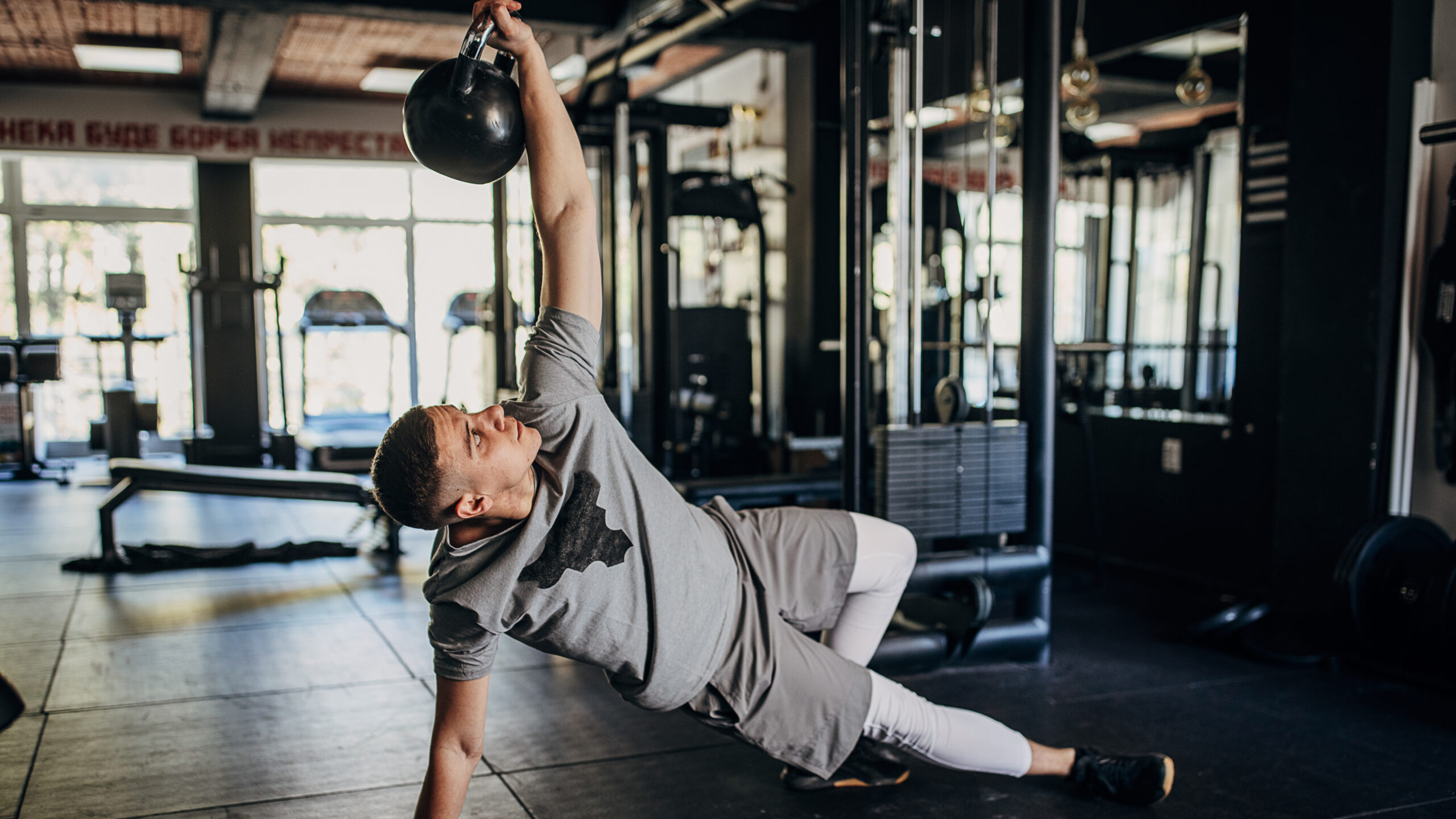 A man performs a Turkish get-up at the gym. He is on the floor, with his body in a straight line, elevated from the ground by his straight right arm and straight right leg. His left leg is bent behind him and he holds a kettlebell straight up in his left hand.