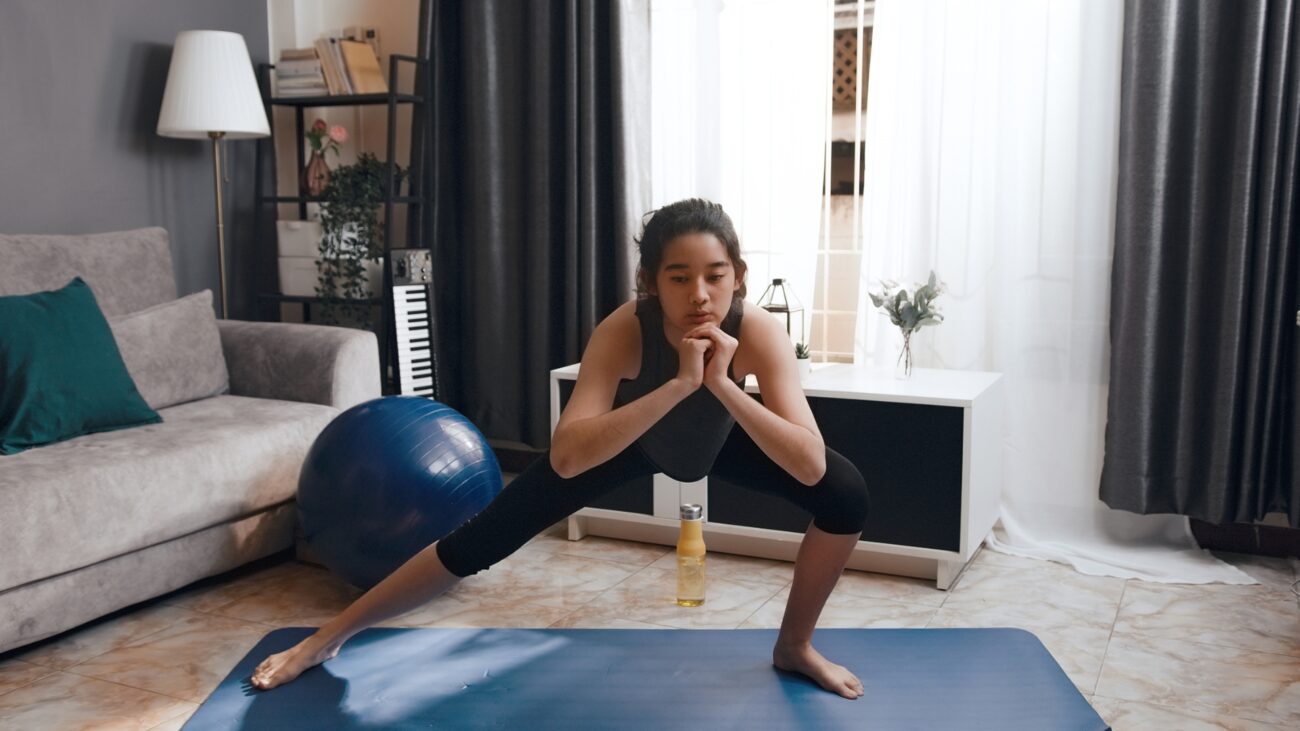 woman doing a side lunge in a living room setting in front of the camera on a blue exercise mat. there's a sofa, window and large exercise ball behind her.