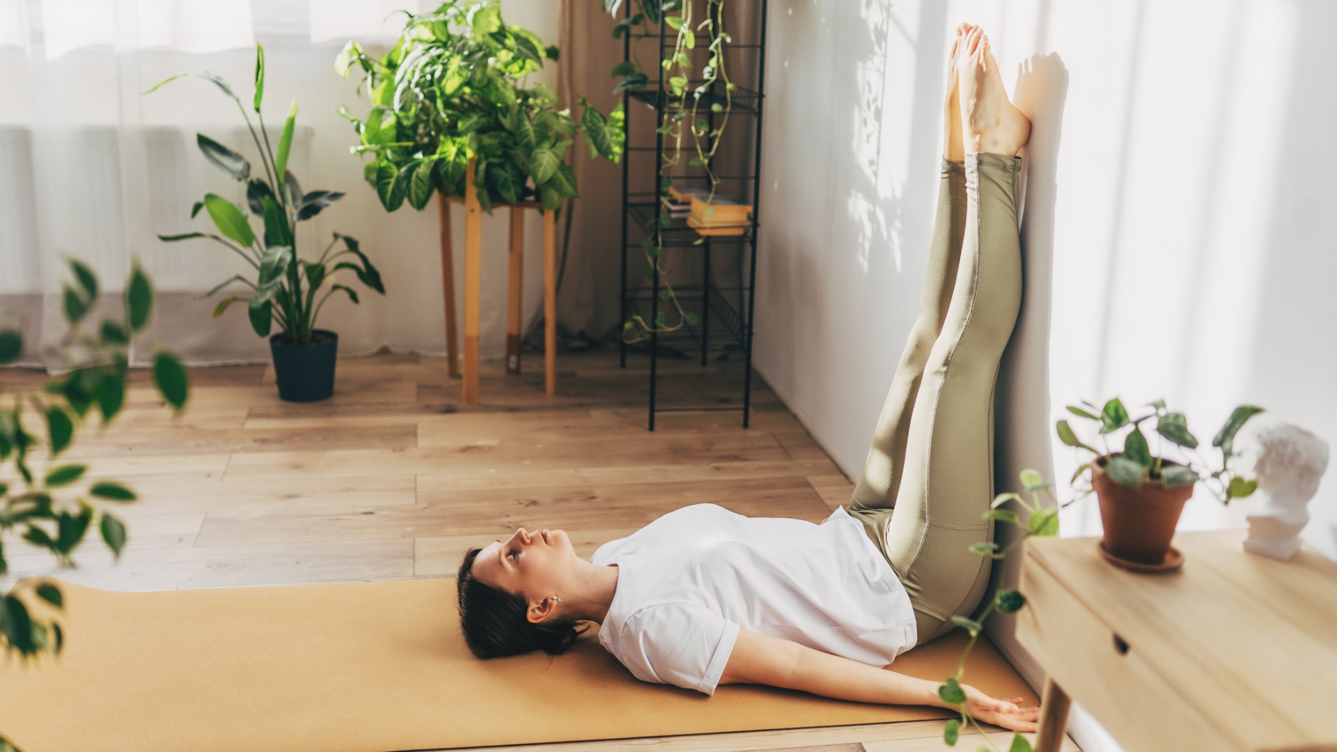 woman wearing white tshirt and khaki leggings on a pale orange yoga mat and wooden floor with her legs vertically up a wall. there are plants around her.