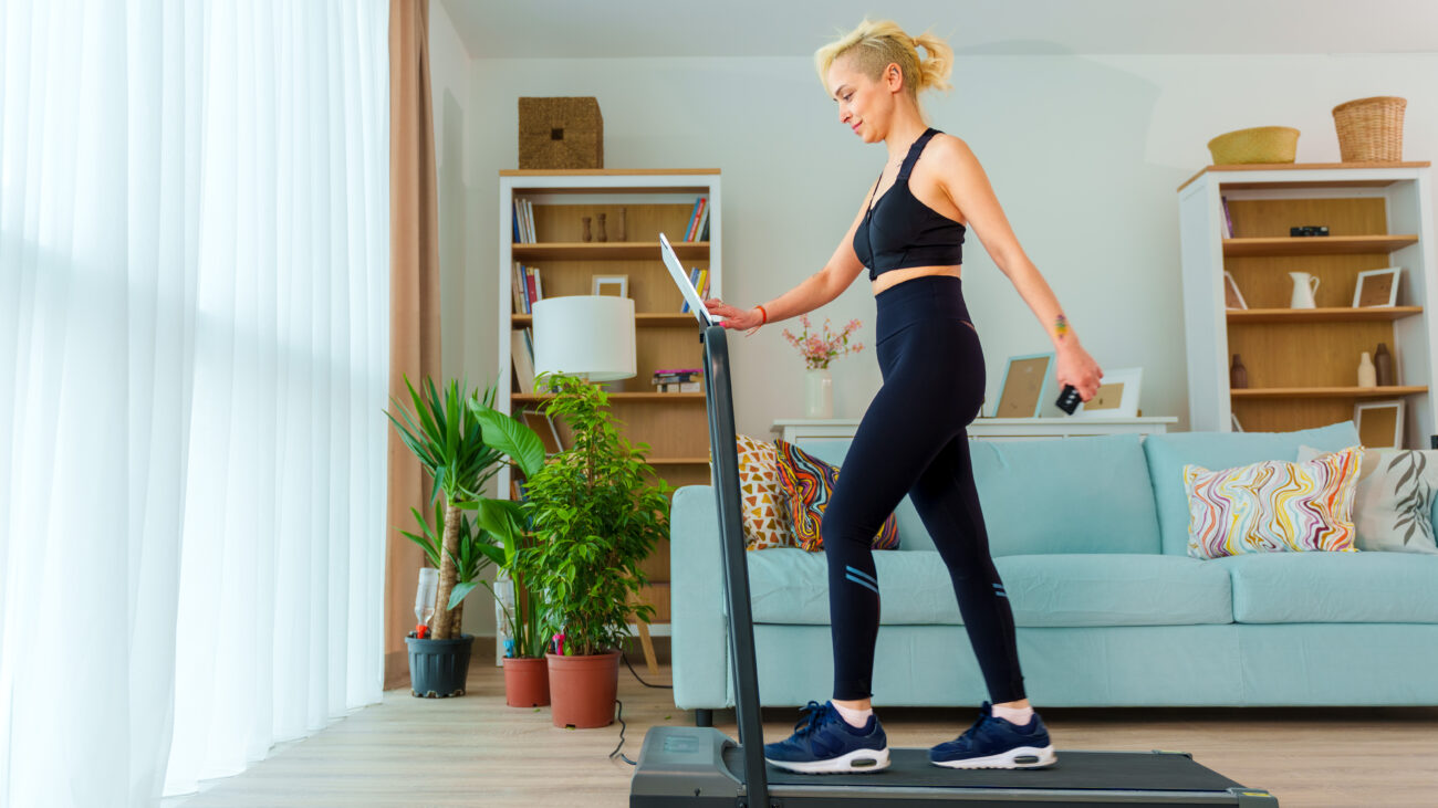 A woman walks on on a treadmill at home in her living room, wearing leggings, a sports bra and sneakers. Behind her is a couch, bookshelf and a few potted plants.