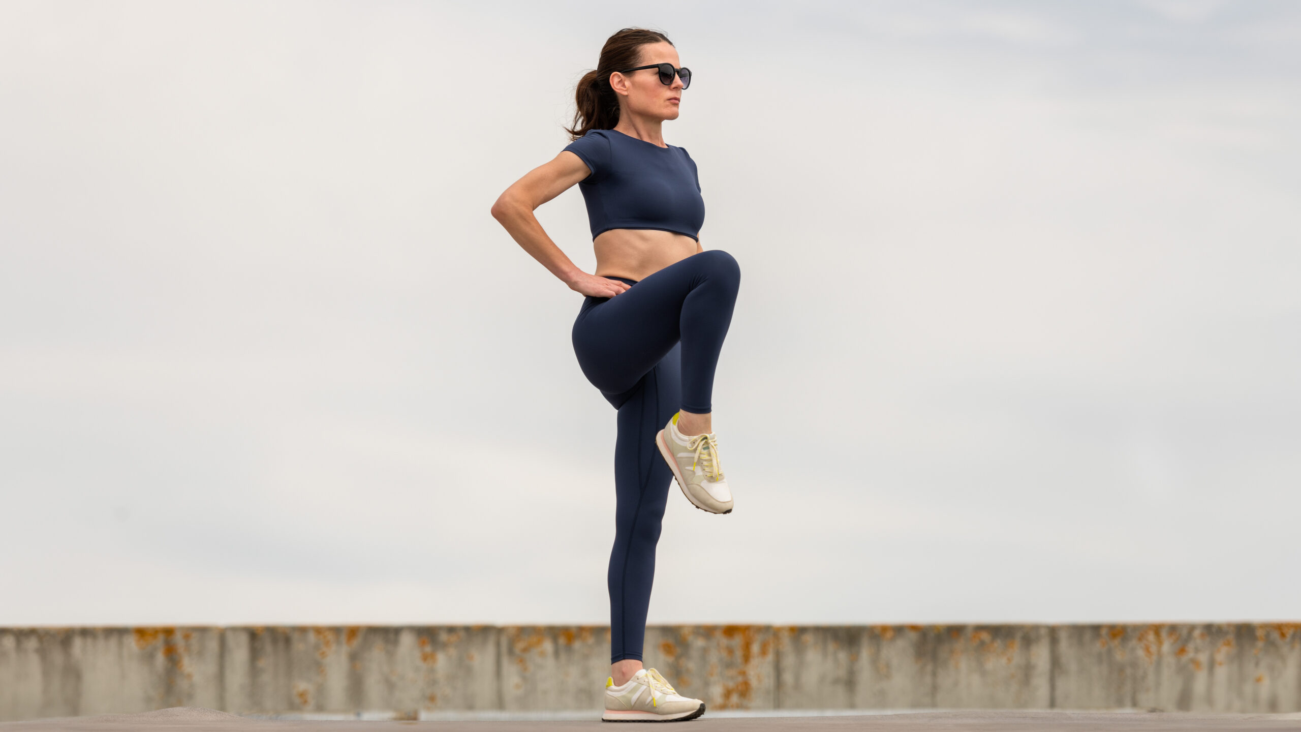 A woman stands with her hands on her hips and one leg raised to hip height. She wears a sporty crop top, leggings and sneakers.