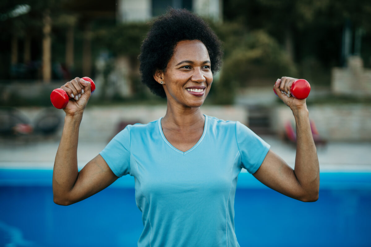A woman stands outside holding a pair of light dumbbells in her hands at shoulder height and smiling.
