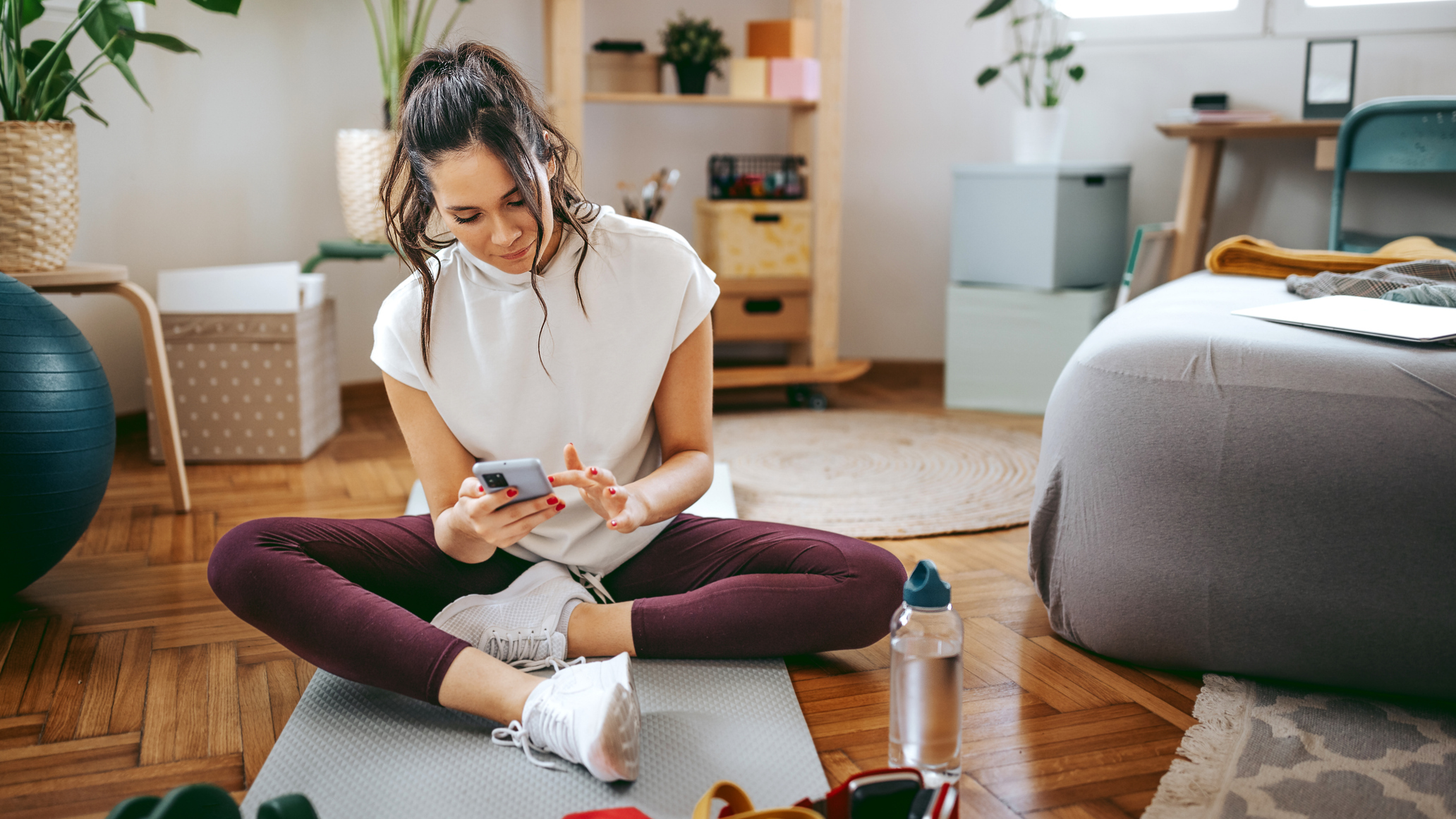 Woman in sportswear sits cross-legged on exercise mat in domestic setting looking at smartphone, with a bottle of water and other exercise equipment in front of her