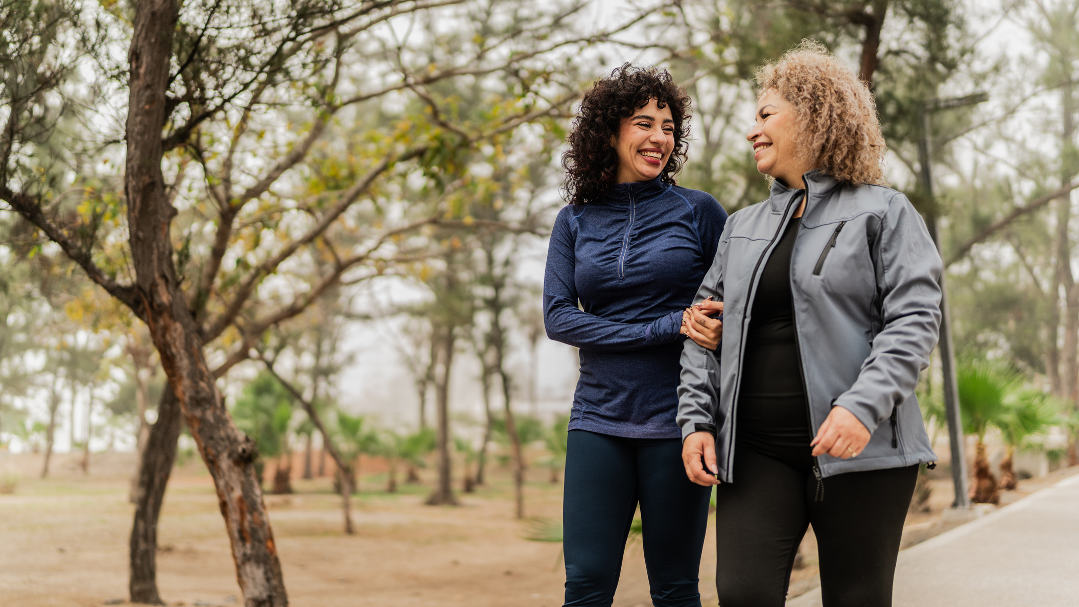 Two women in workout tracksuits walking arm in arm in a park smiling at eachother