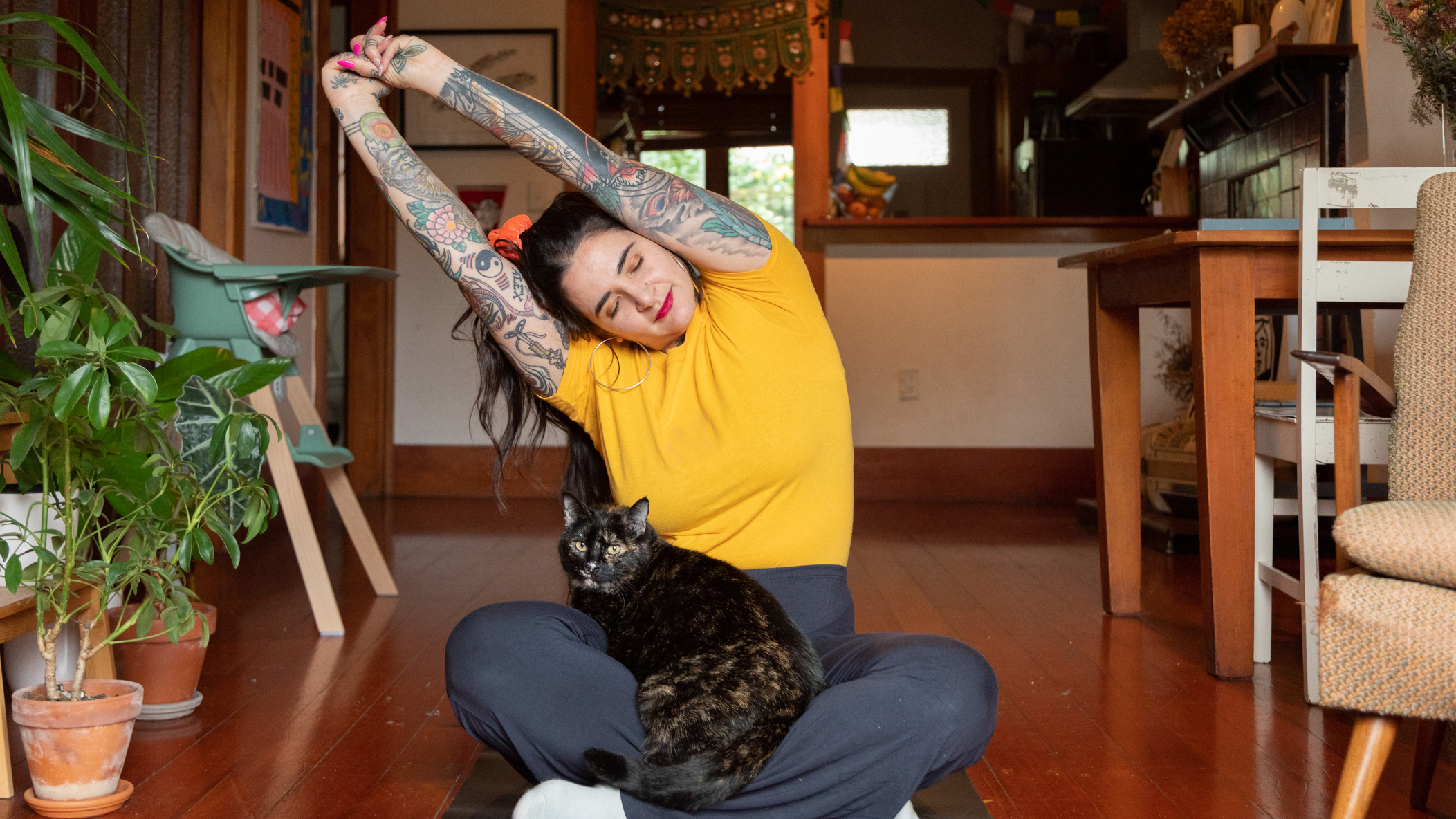 Woman sits cross-legged on floor with a cat on her lap. She stretches her arms overhead and to the side. She is in a domestic setting.